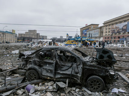A city square is littered with rubble. A burned-out car is in the foreground. People with yellow armbands are in the background lined up outside of a large tent.