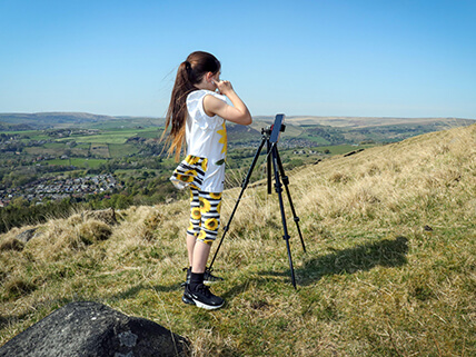 A girl is standing on a hillwith a village in the background. She has a phone set up on a tripod to record herself.